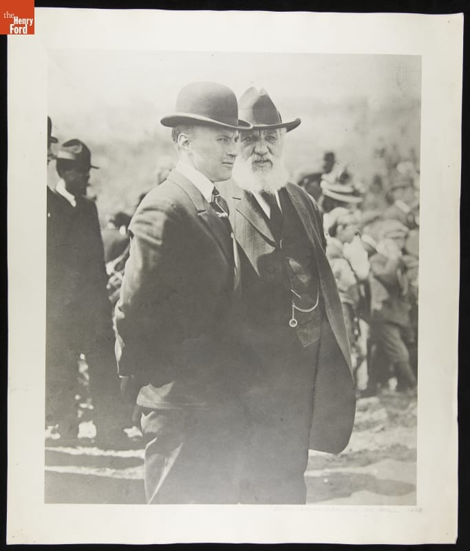 Black-and-white photo of two men in suits and hats talking to each other among a crowd