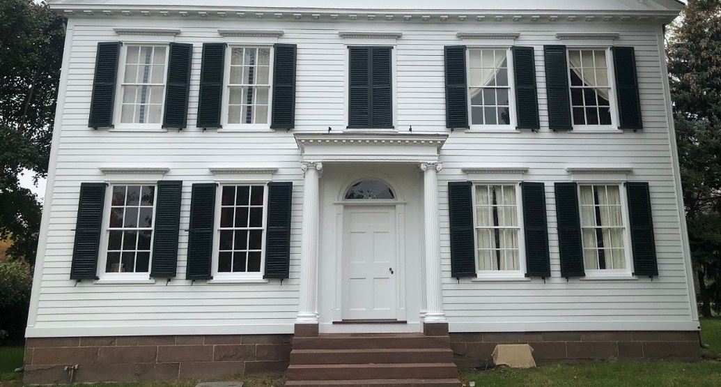Front façade of a white house with symmetrical windows with black shutters, and closed black shutters above the front door