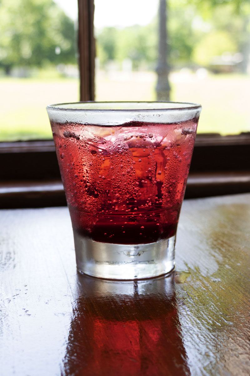 Red-colored drink with ice in glass sitting on wooden table by window