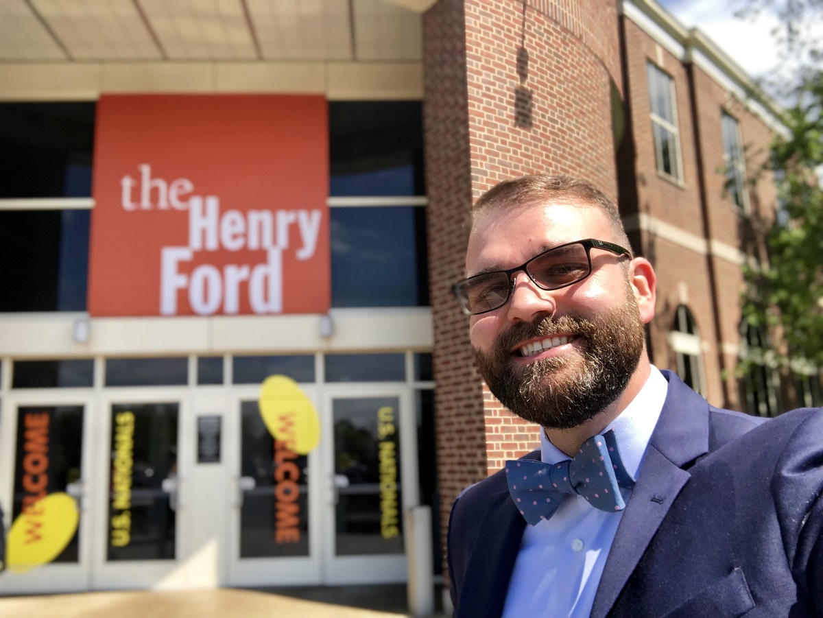Teacher Spencer Kiper Man in blue suit and bow tie takes a selfie in front of a building with large logo reading "The Henry Ford"