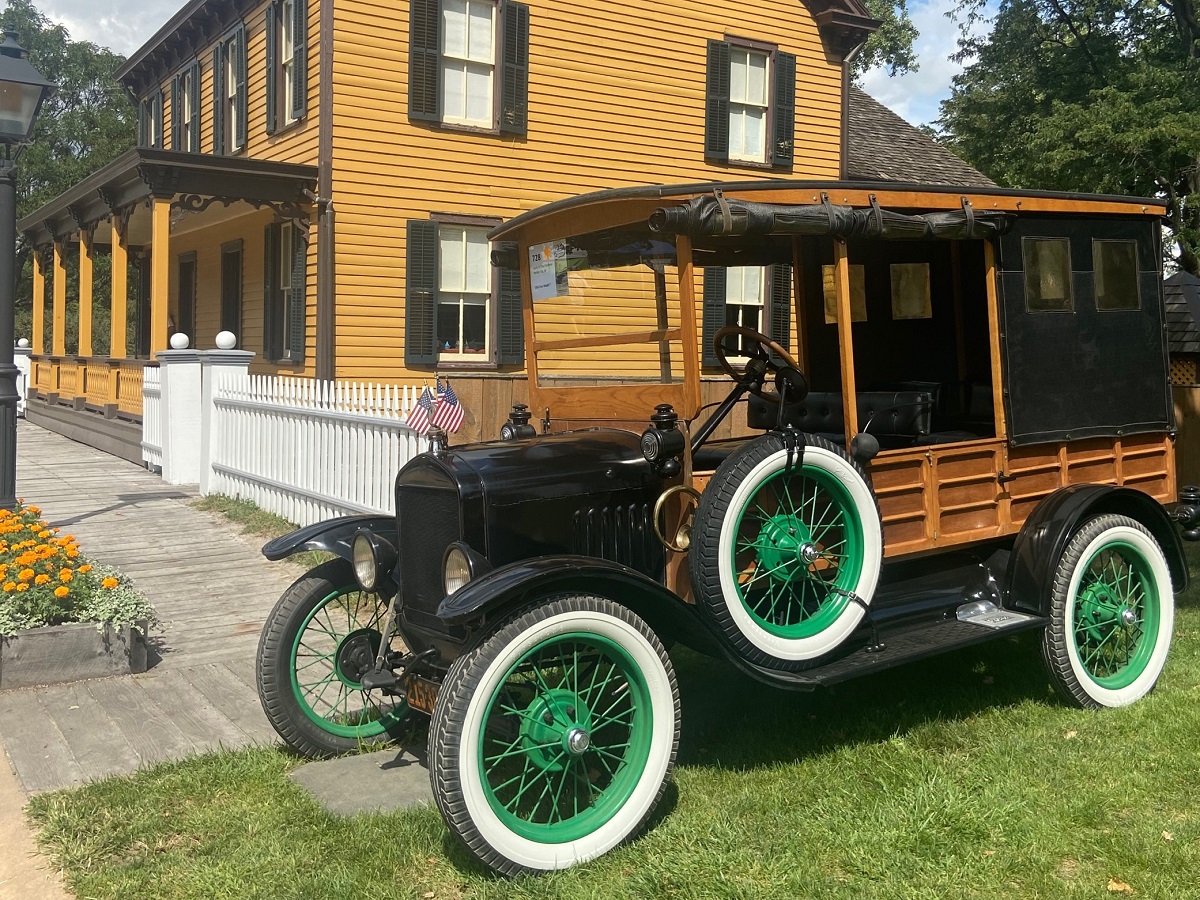 Old-fashioned truck with wooden sides and whitewall tires with green rims, parked in front of a mustard-colored wooden building
