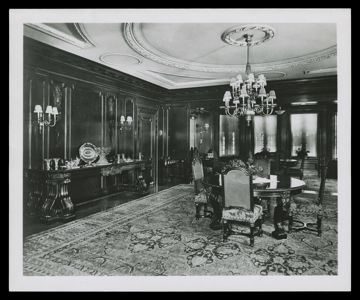 Room interior with round dining table and four chairs, oriental rug, and ornately carved wooden walls and plastered ceiling