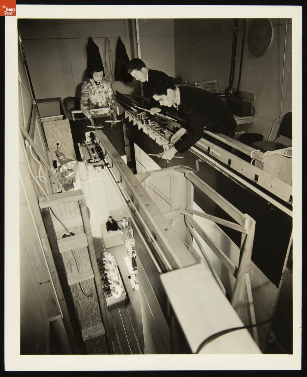 Three people on an elevated balcony or catwalk hold wooden crosspieces supporting puppets below
