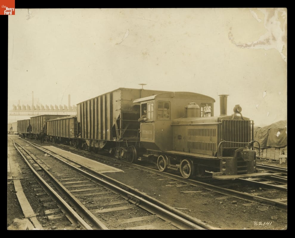 Black-and-white photo of small locomotive pulling rail cars on railroad tracks