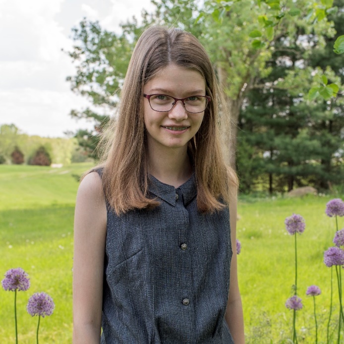 Young woman with blonde hair and gray sleeveless top stands among flowers with grass and trees behind her