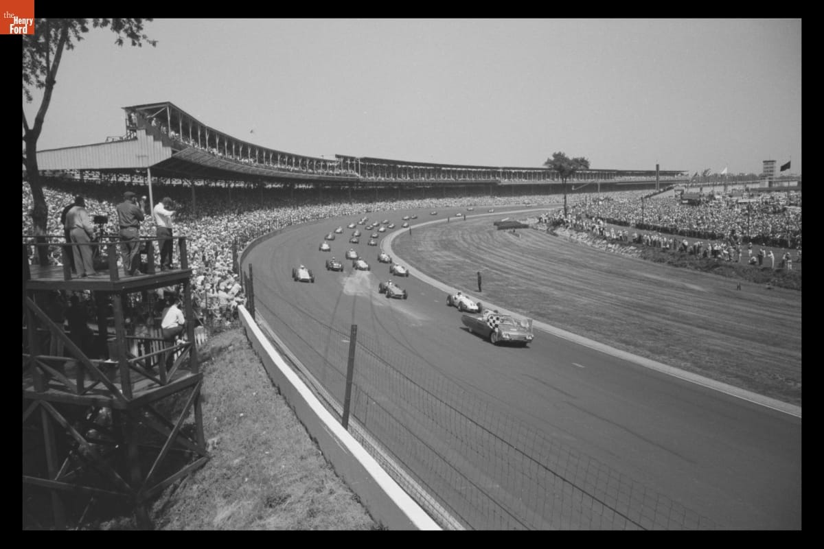 Black-and-white photo of many cars on race track, with grandstands full of fans on both sides of the track