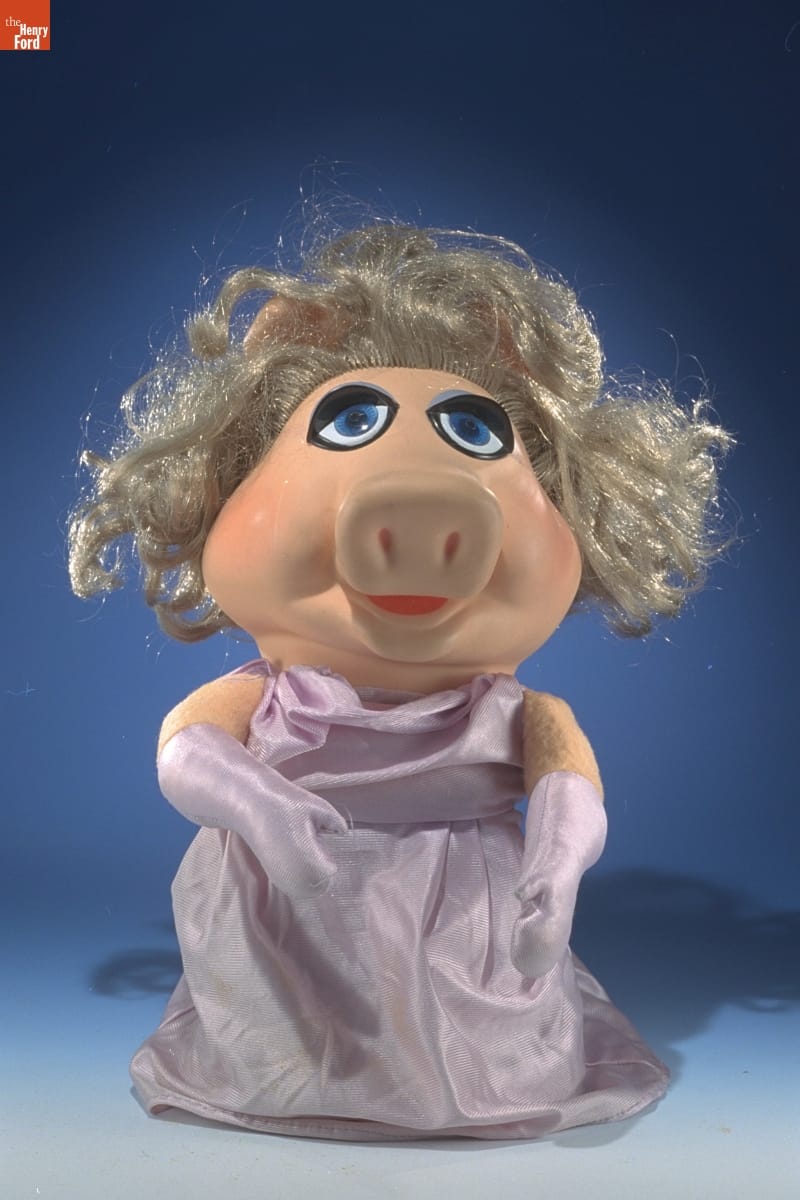 Puppet of pig with blonde hair wearing purple dress and purple shoulder-length gloves
