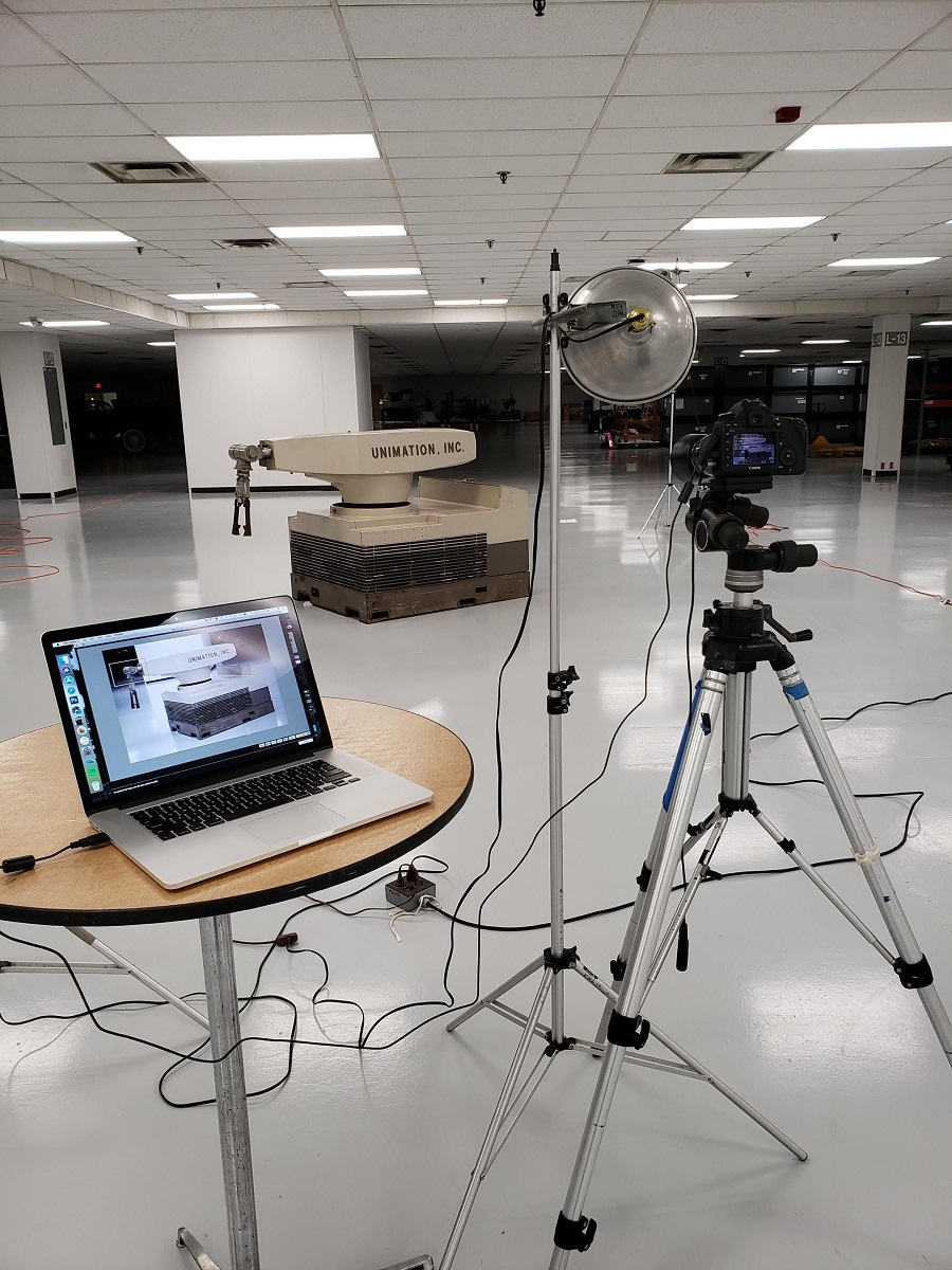 Large, squat robot  with swing arm in the middle of a  large space; photo on tripod and tethered laptop computer on table in the foreground