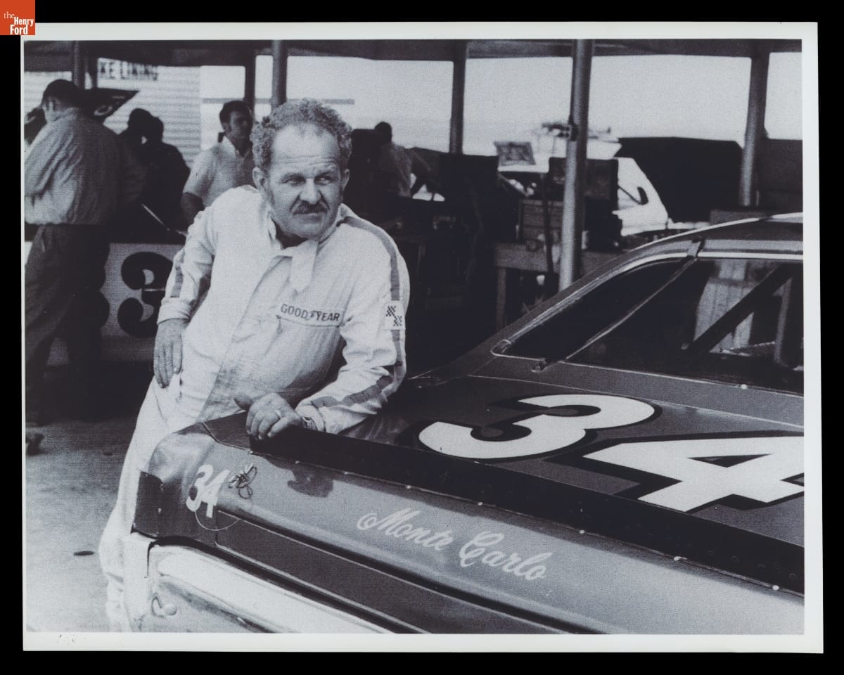 Man in jumpsuit with short curly hair and mustache leans against back of race car, with other race cars, people, and equipment in the background