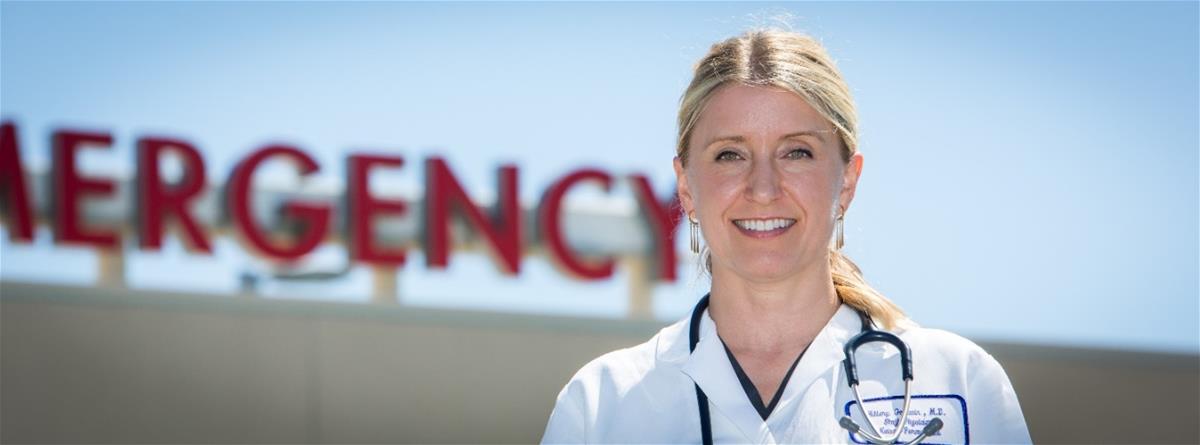 Light-skinned blonde woman in medical coat with stethoscope around neck stands in front of sign reading "EMERGENCY"