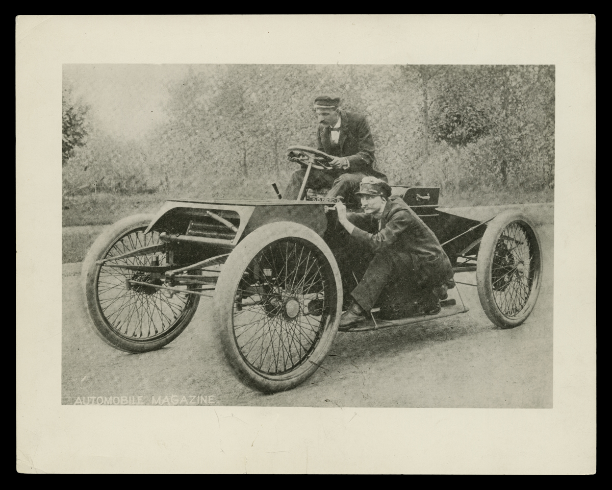 Early open automobile on street with one man behind wheel and another crouching on running board