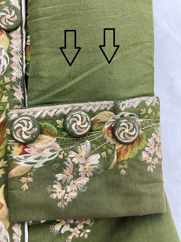 Markings from original button placement are visible above top of cuff Detail of moss-green coat sleeve with elaborate embroidery and embroidery-covered buttons; two arrows point to two slight picks in the fabric above the cuff