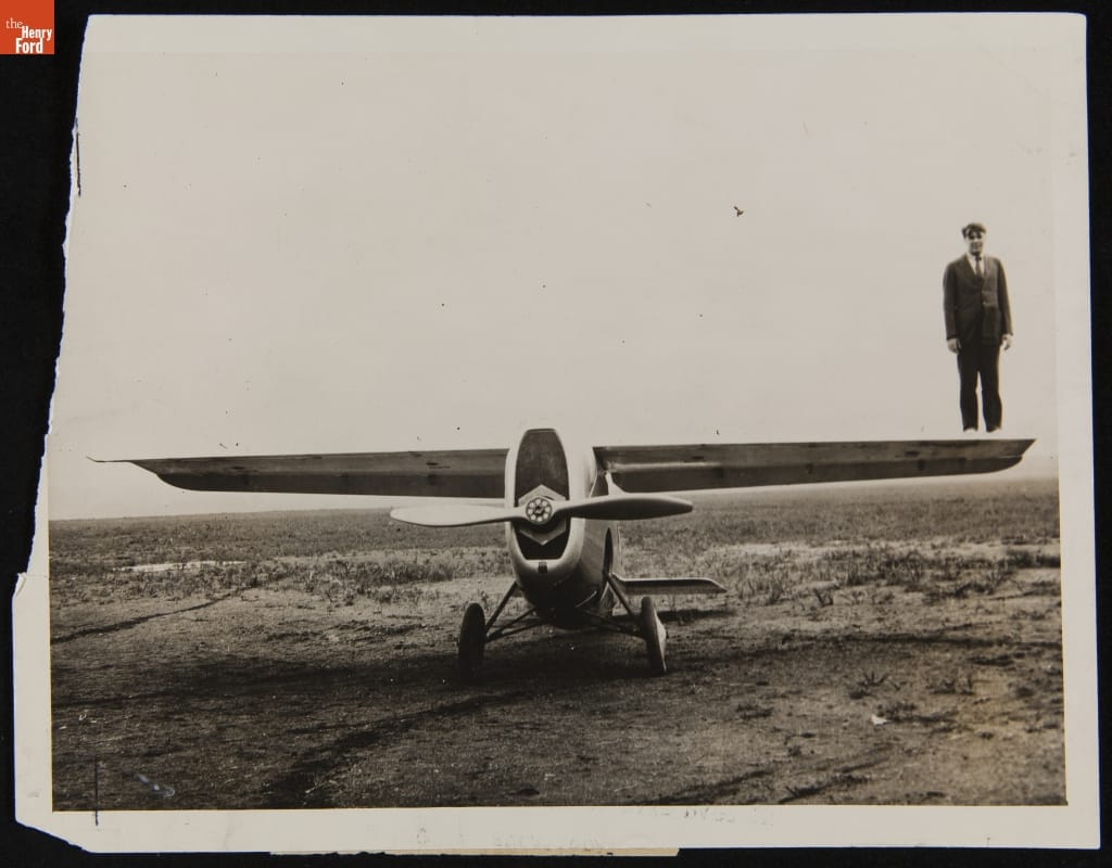 Black-and-white photo of man standing on top of end of wing of small airplane parked in large open field