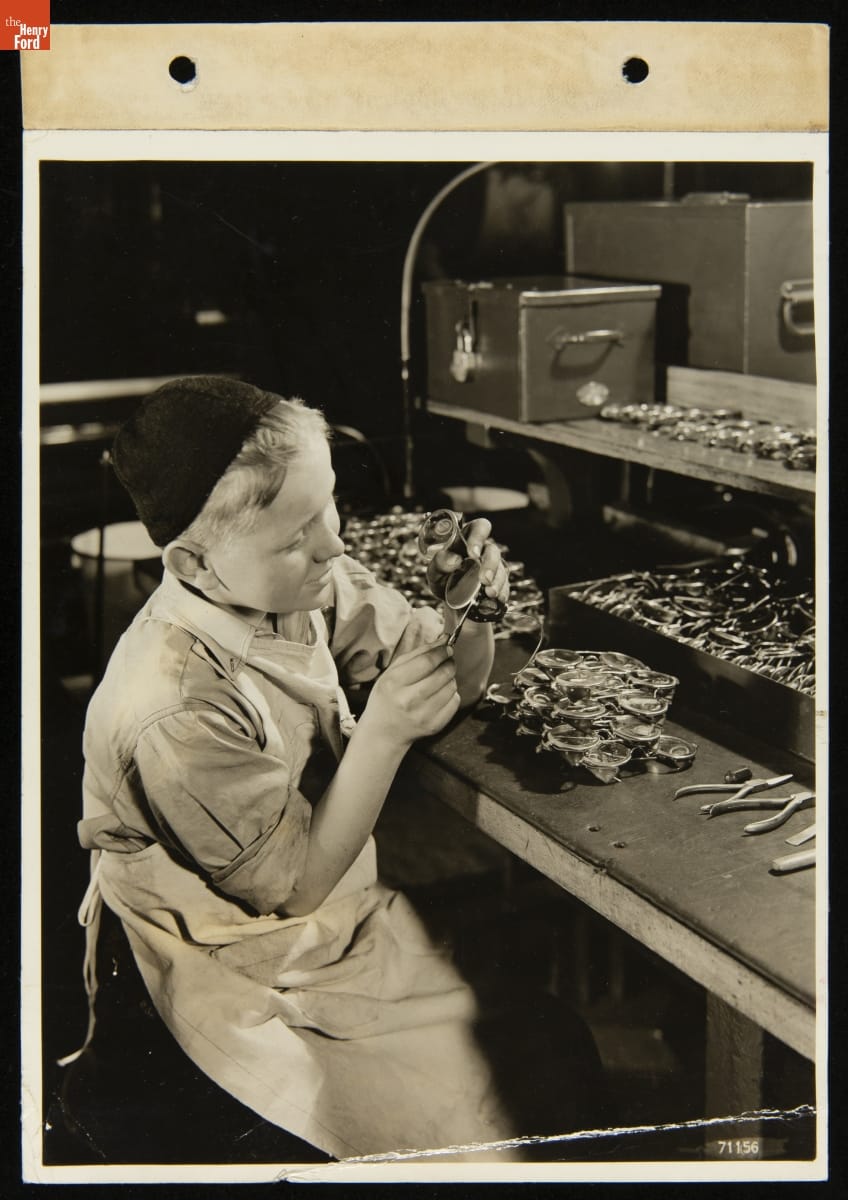 Boy wearing apron and cap holds tool to a pair of goggles; a table nearby holds many additional pairs of goggles