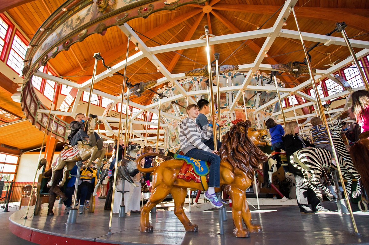 Low-angle photo of a carousel in a wooden structure with children riding carved animals