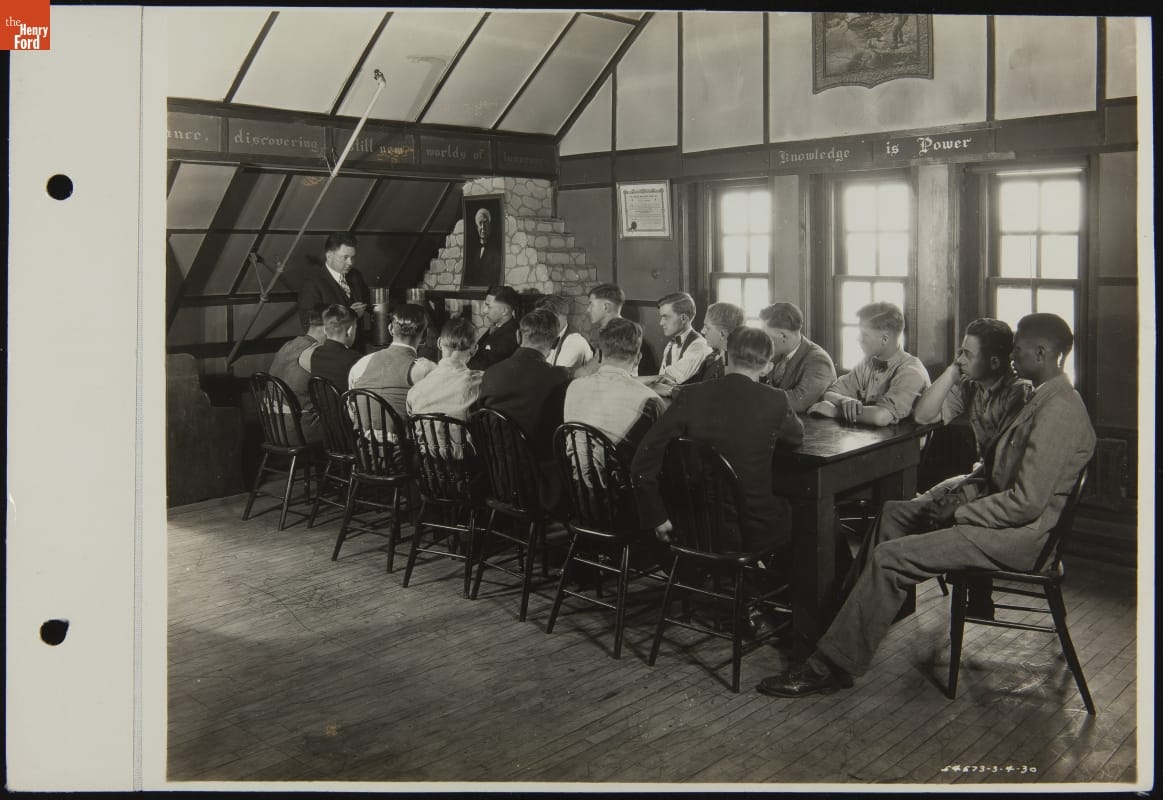 Young men sit around a long table looking at a man standing at one end 