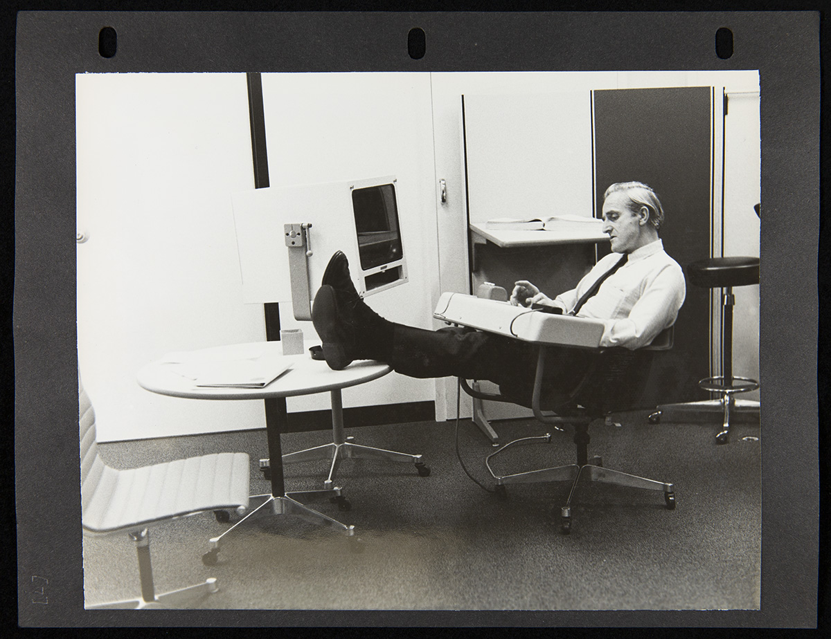 Page from Report on the SRI Fall Joint Computer Conference, December 27, 1968 Page from three-ring binder containing photo of man sits in chair with attached console with feet up on a nearby table that also holds what appears to be a large monitor; office furniture around him