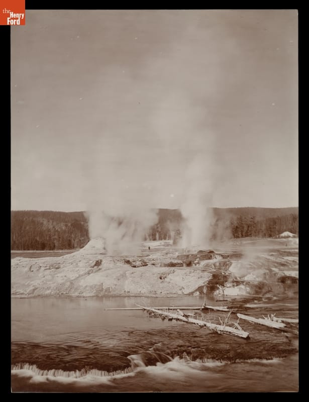 Geysers at Yellowstone National Park, "The Grand Group," 1870-1880 Black-and-white photo of geysers erupting steam and/or water into the air in a rocky landscape with a lake or river in the foreground and forest in the background
