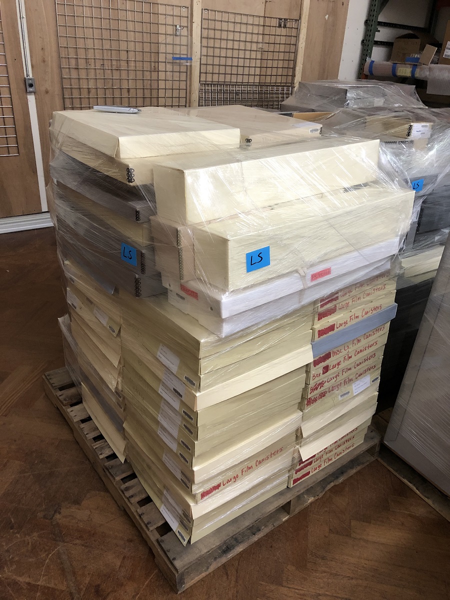 Pallet containing many wide, shallow boxes, all wrapped in clear plastic wrap, sitting in a storage area among other similarly packaged material