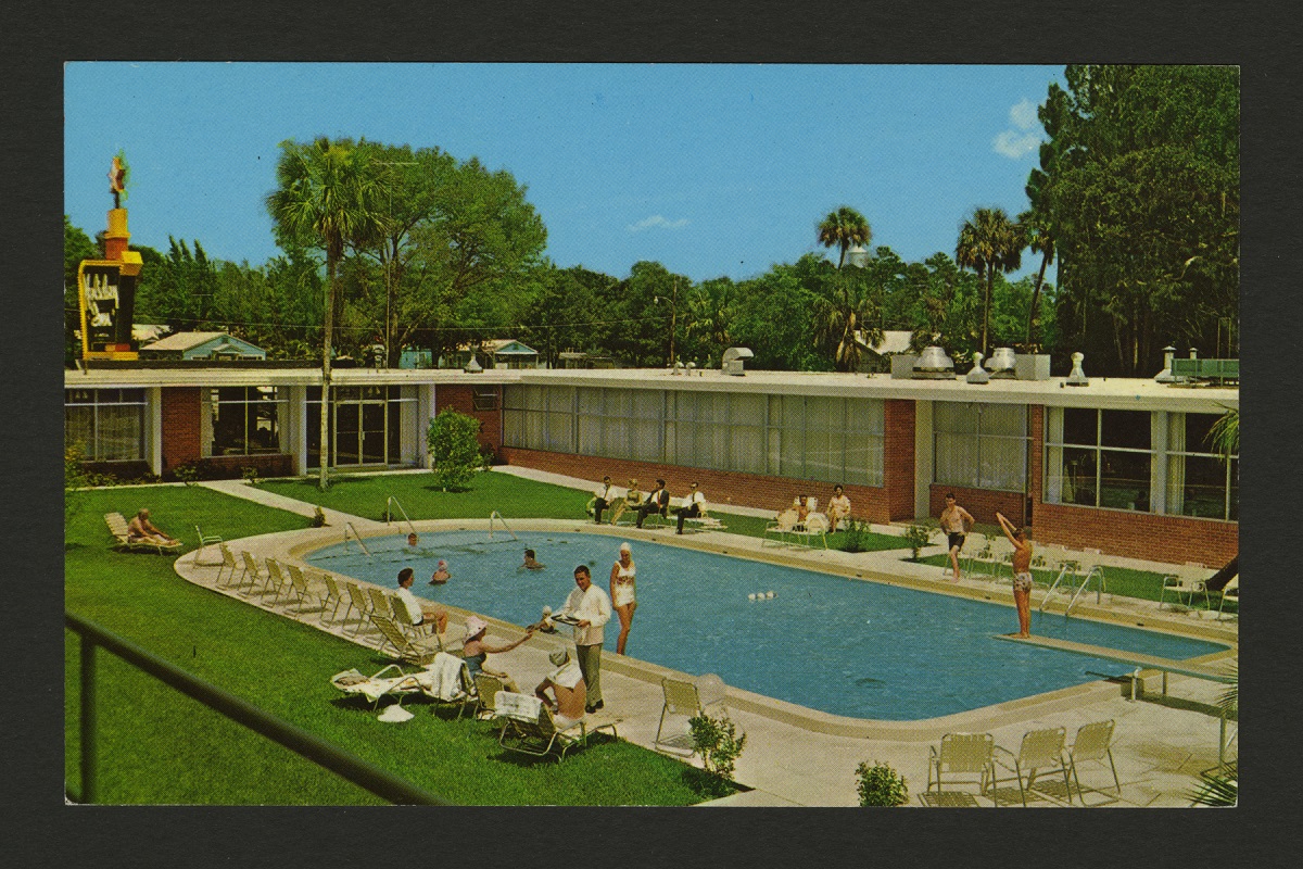 L-shaped low brick building bordering grassy area containing swimming pool with people in and around it