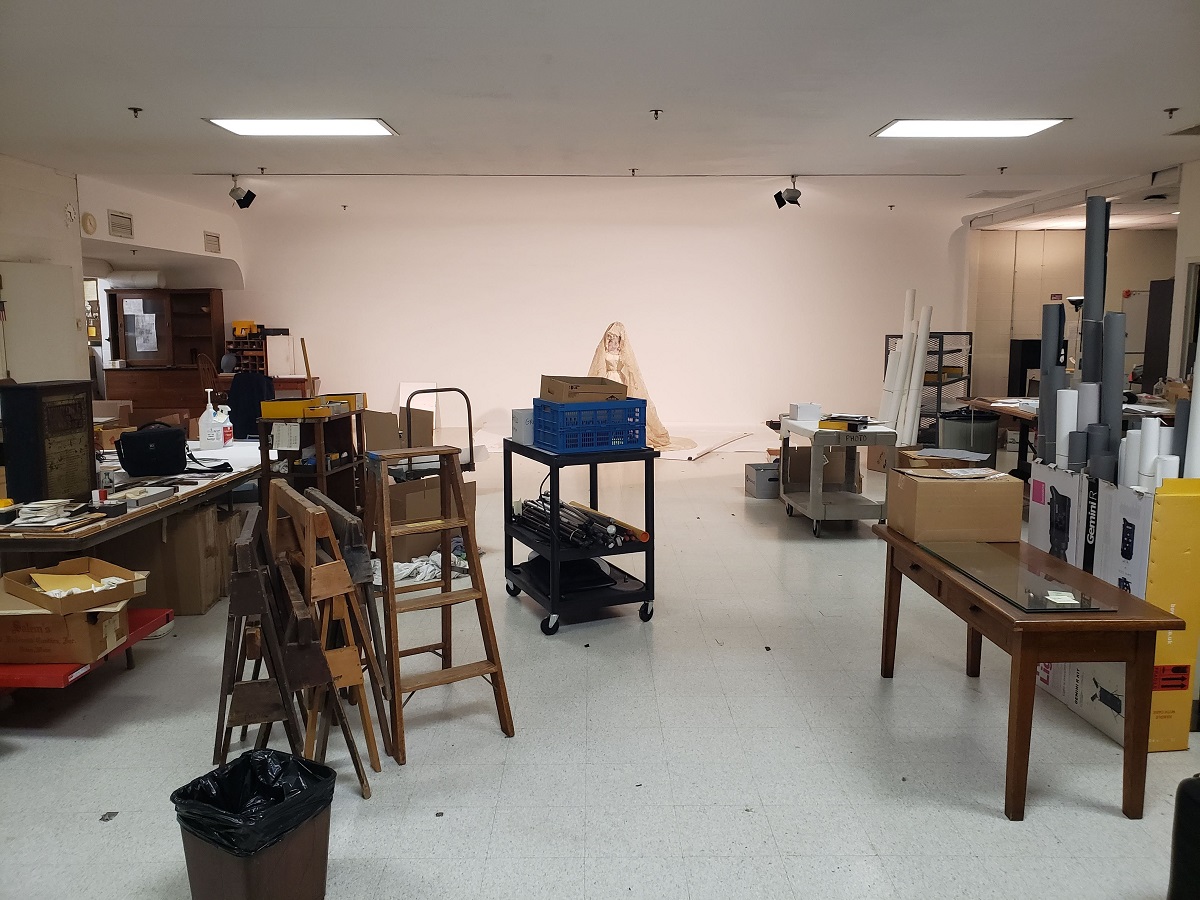 Photographing the final object in the old studio Room filled with furniture, stacked ladders, carts, sawhorses, and a wedding dress on a mannequin in the background