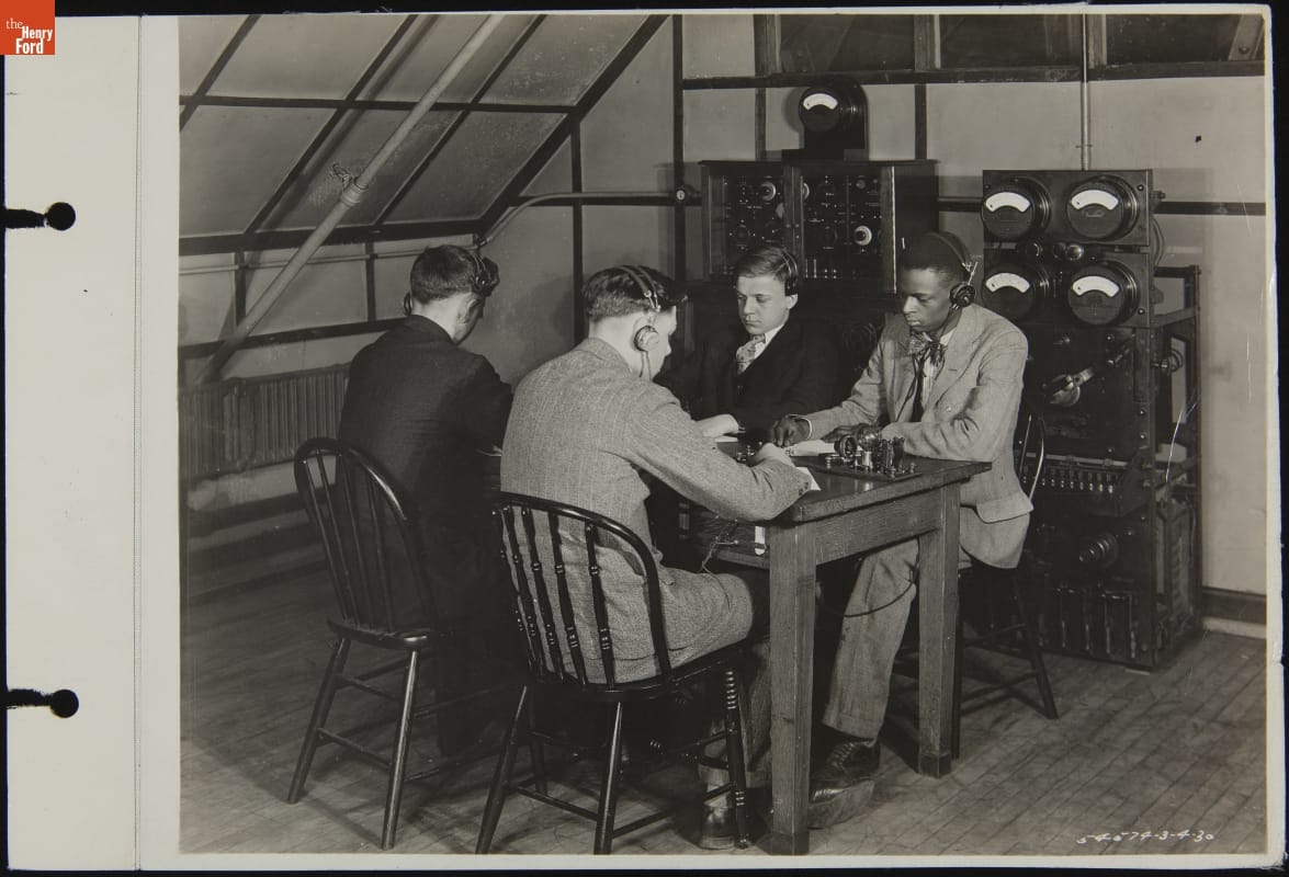 Four men in suits wearing headphones sit at a table with equipment on the table and along the wall behind them