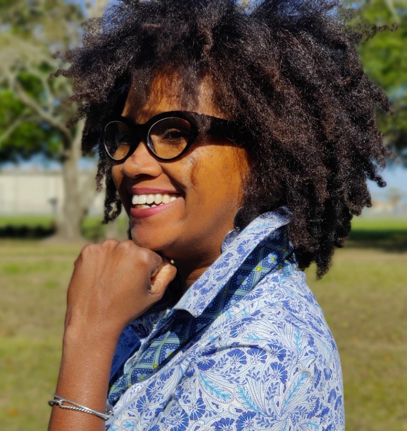 Dark-skinned woman with natural hair, glasses, and wearing a blue print shirt holds a fist to her chin and smiles