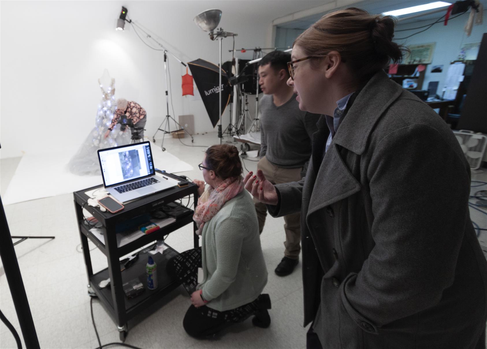 The dress in the studio getting ready for its close-up with curator Kristen Gallerneaux and conservators Fran Faile and Cuong Nguyen assisting. Three people around a computer on a cart, with photography equipment and a woman adjusting a dress on a dress form in the background