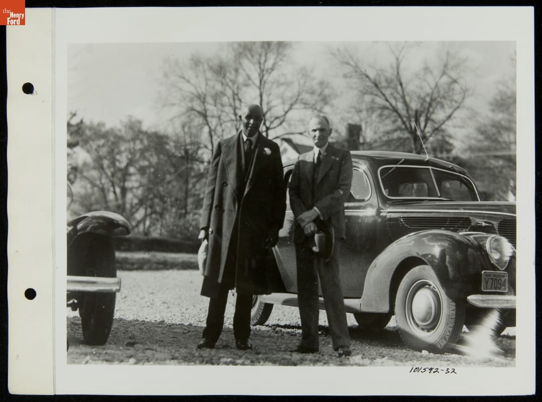 Two men, one Black and one white, pose in front of a car