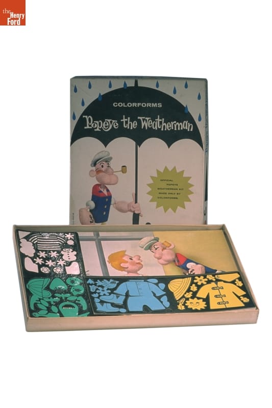 Open box with lid standing on end behind it; box contains colorful pieces on a sheet; box lid contains image of sailor Popeye character with umbrella and text