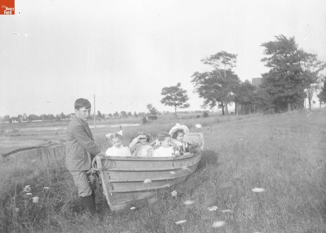 Young boy pulls at the front of a wooden rowboat in a grassy field, while four girls sit inside