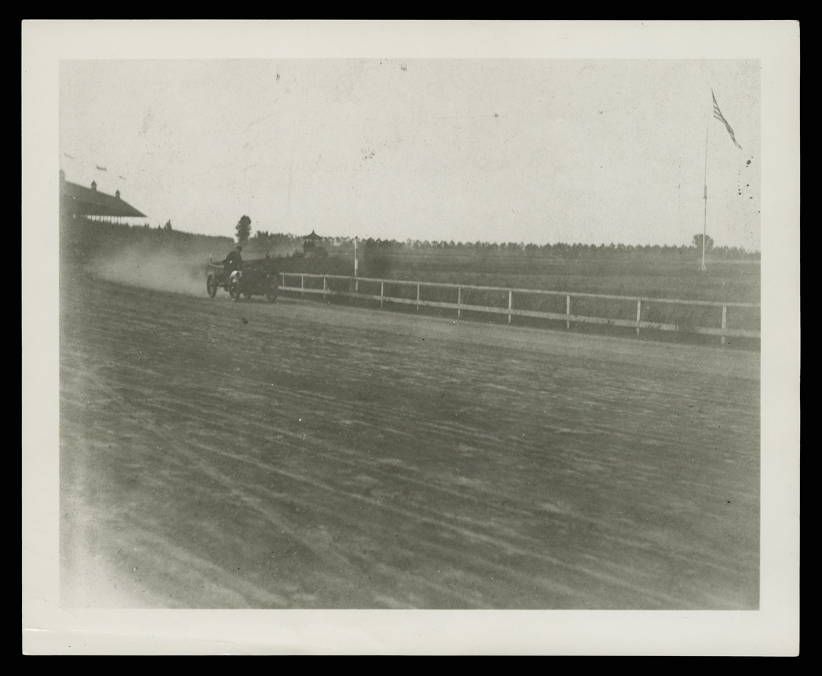 Early minimal race car in the mid-distance on a dirt track with a fence and grass behind it