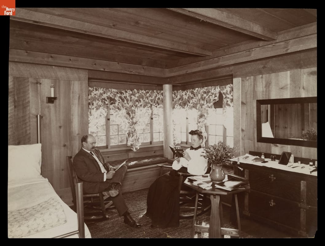 A Typical Bed Chamber in the Old Faithful Inn, Yellowstone National Park, circa 1905 Man and woman sit reading in rocking chairs in a rustic bedroom with wooden walls