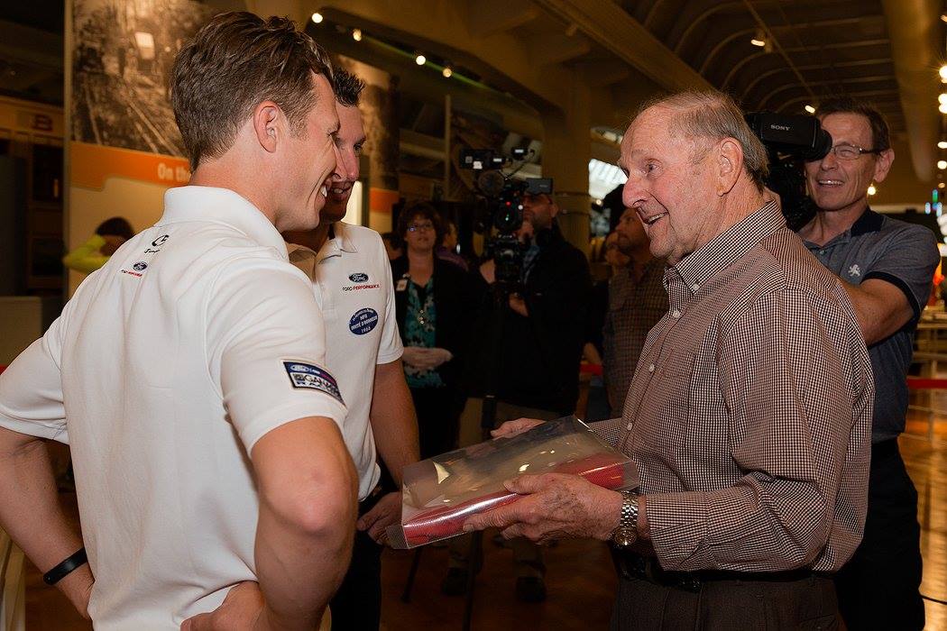 Smiling man hands a package to two other men, while people look on and camerapeople film