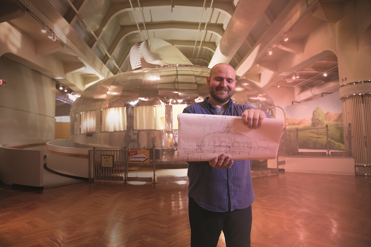 Man stands in large building with round silver metal structure behind him, holding a drawing of the same structure