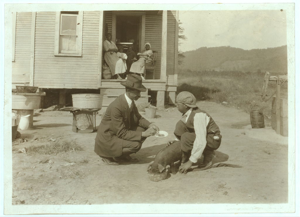 Man in suit and hat kneels near youth and pig eating from dish in a dirt yard; women watch from a porch behind them