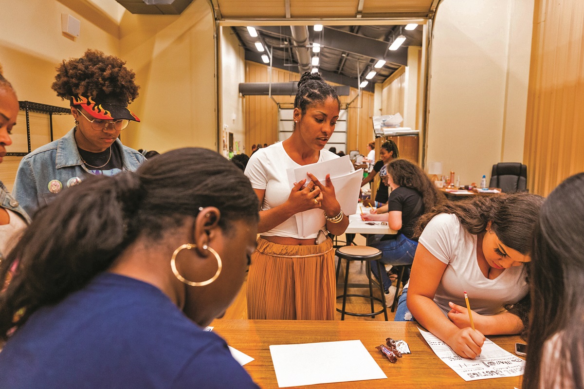 A group of young people write on paper around a table, with other groups working at other tables in the background
