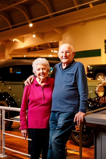 Man in blue shirt and blue jeans leans on barrier with his arm around woman in pink shirt and black pants in front of car display