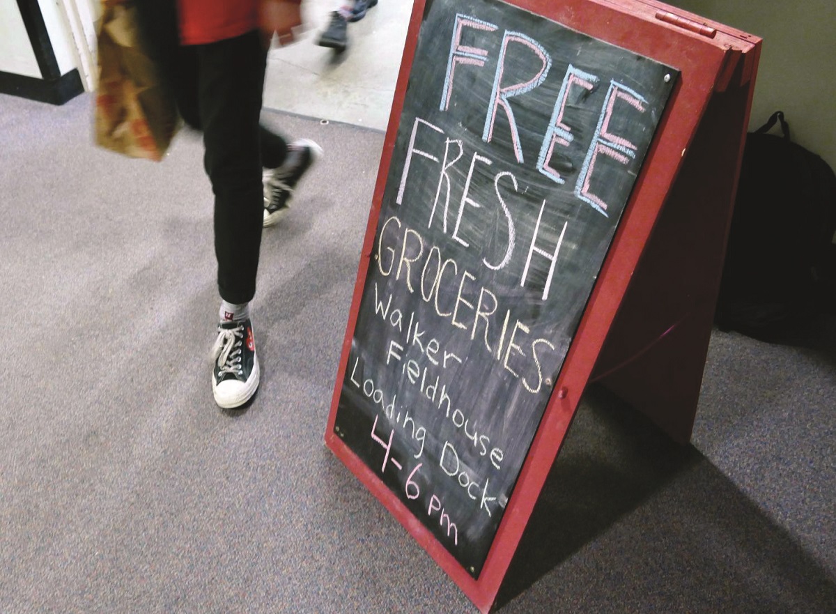 Standee-type blackboard sign reading "Free fresh groceries: Walker Fieldhouse Loading Dock 4-6 PM" with person walking by it holding a paper grocery bag