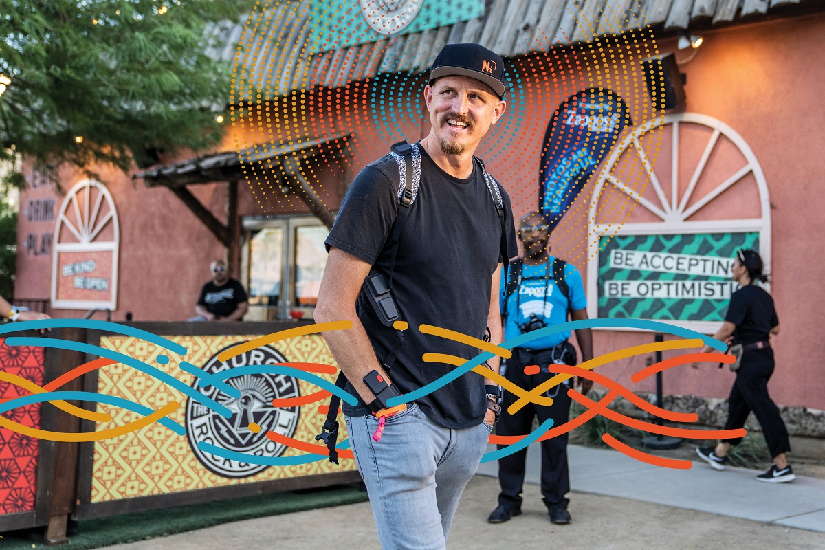 Man in black t-shirt, jeans, baseball cap, and backpack stands in a plaza with a few other people and red building behind him