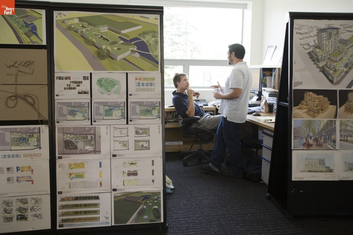 Two people, one seated and one standing, talk in a sunny office cubicle; drawings and renderings of buildings hang on outside walls