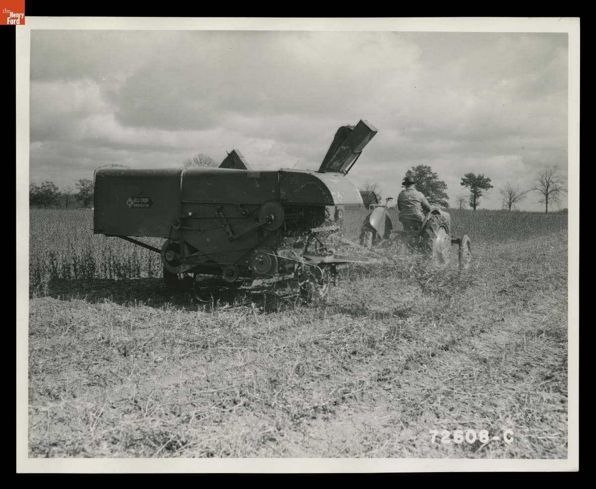Black-and-white photo of person on tractor pulling a piece of agricultural equipment behind them through a field
