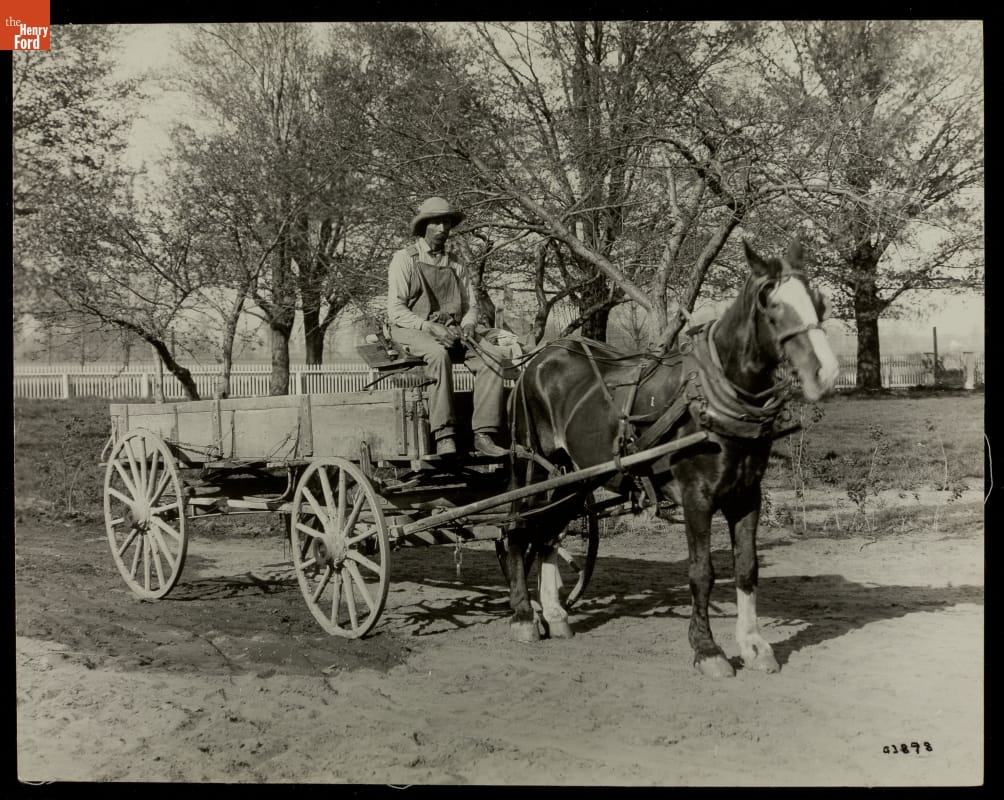 Man in overalls and hat sits on driver's seat of an open wagon hitched to a horse