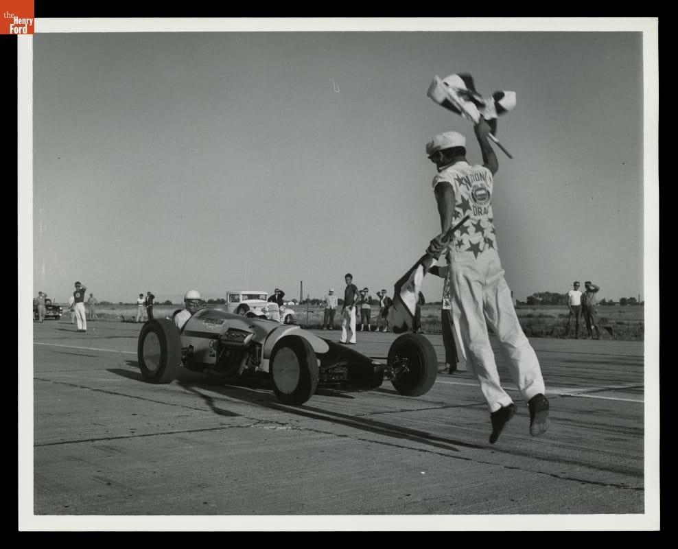Person holding a checkered flag in each hand in mid-jump in front of a race car on a track