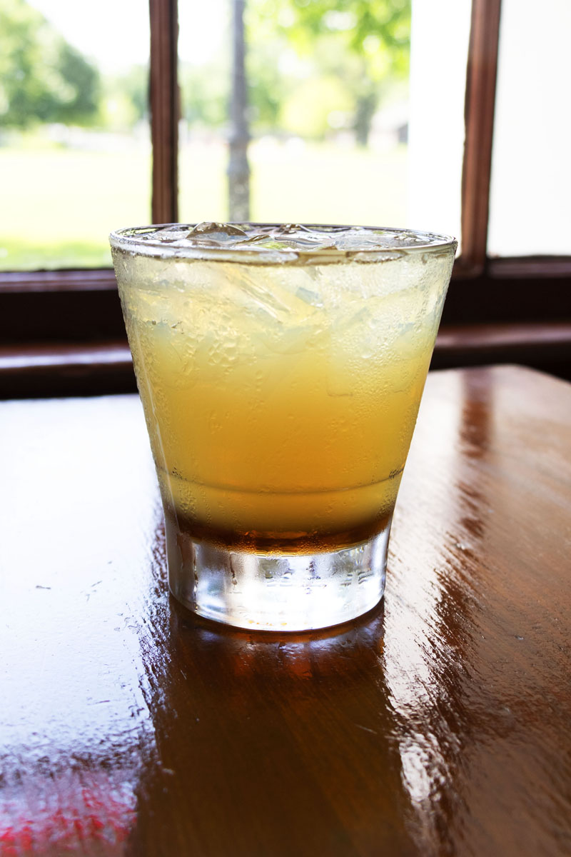 Honey-colored drink in clear glass with ice, sitting on wooden table in front of window