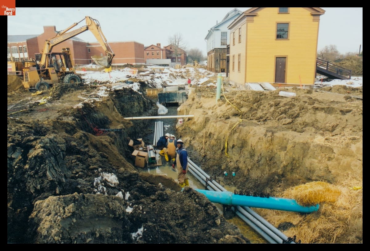 Workers Laying Conduit in Greenfield Village during Infrastructure Restoration, January 2003 Three men with pipes in muddy trench; construction equipment and buildings nearby