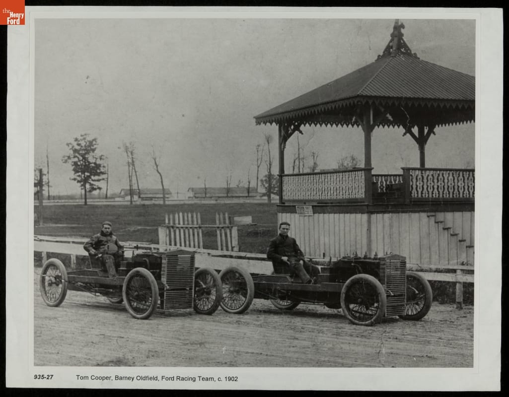 Two men pose at the wheel of two very minimal open early race cars on a track next to a covered pavilion