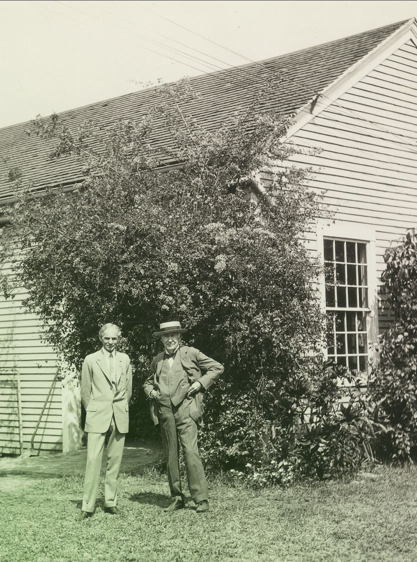 Two men in suits, one wearing a hat, stand in front of a wooden building partially obscured by vegetation