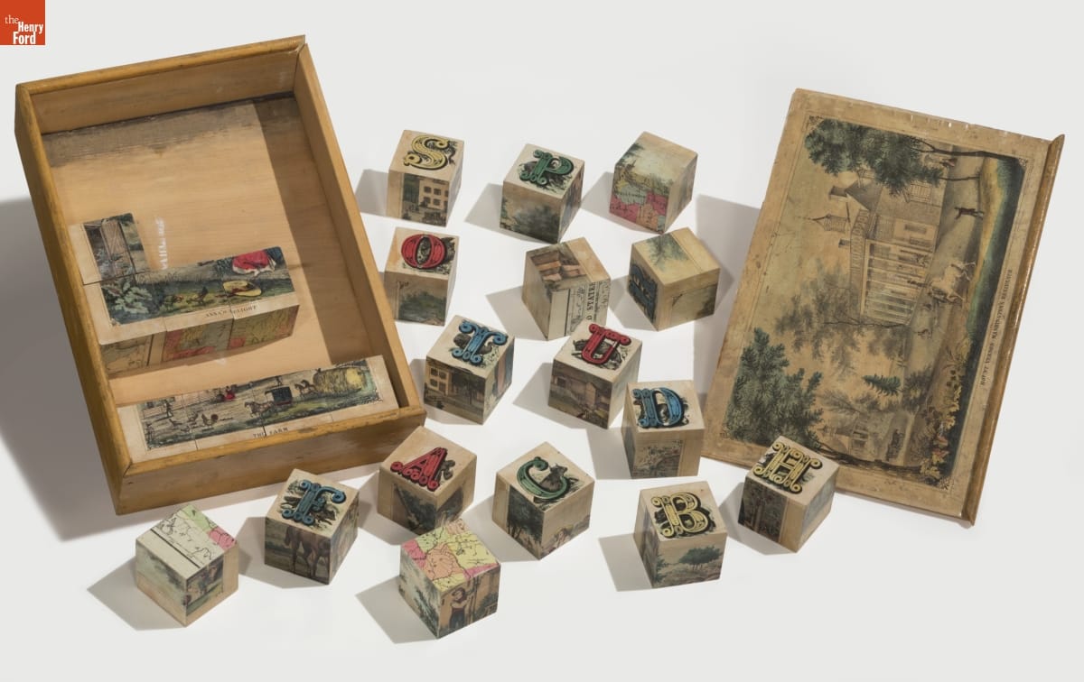 Wooden box with some blocks in it and other blocks scattered around; top of box contains image of house and grounds