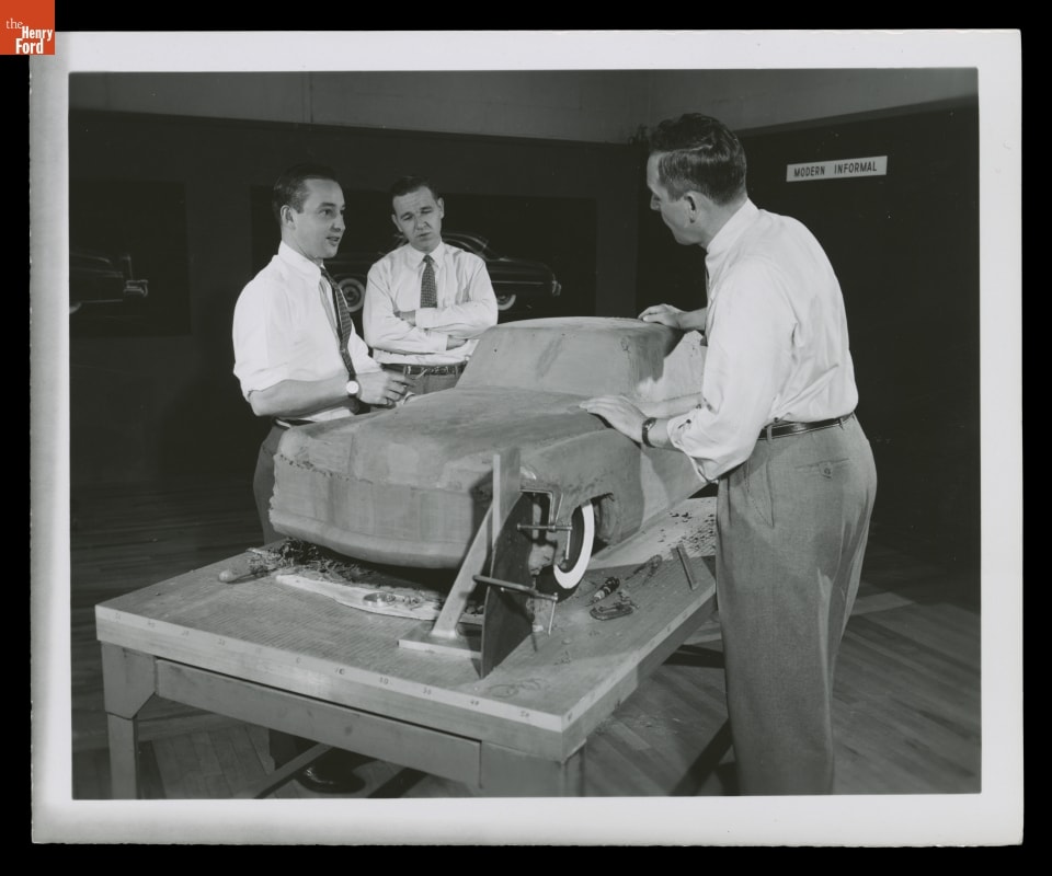 Three men standing around clay model of car on table